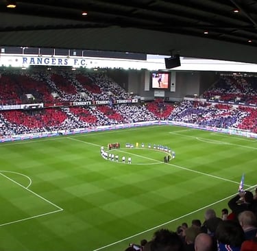 Innenansicht des Ibrox Stadium mit grünem Spielfeld und blauen Sitzreihen.