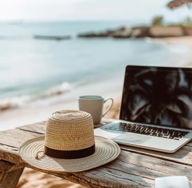 A laptop computer on a table with a hat and coffee mug, overlooking a beautiful morning beach scene 
