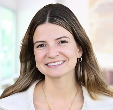 Professional headshot of a smiling young woman with brown hair, gold jewelry, and a white blazer.