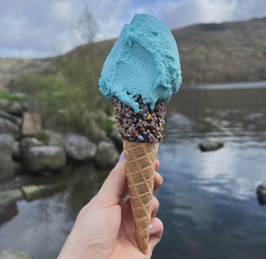 Image is a one scoop sprinkle cone with bubblegum ice cream. View of Llyn Padarn in background.