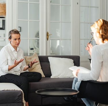 Two women in a therapy session, one gesturing while speaking, the other taking notes