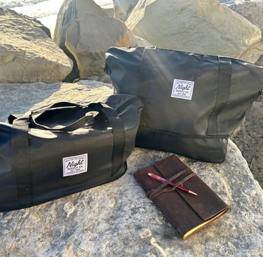 Two black Night Supply Co. duffel bags on rocks beside leather journal and pen.
