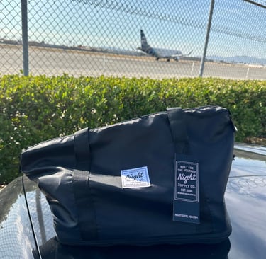 Black Night Supply Co. duffel bag with logo and hang tag, placed near airport fence