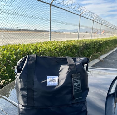 Black Night Supply Co. duffel bag with logo and hang tag, placed near airport fence