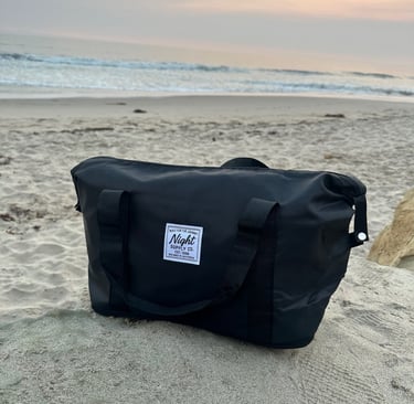 Black travel bag from Night Supply Co. placed on a sandy beach at sunset, with ocean waves and mount
