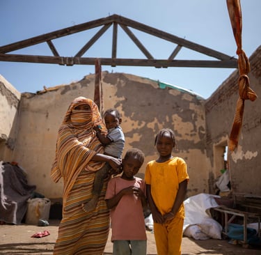 Sudanese woman and children standing inside a roofless home damaged by conflict.
