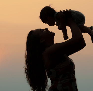 Mother holding child up in the air during sunset, both silhouetted against the sky