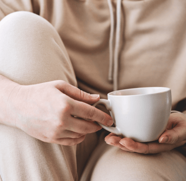 Person in loungewear holding a white mug while sitting with knees up on a sofa