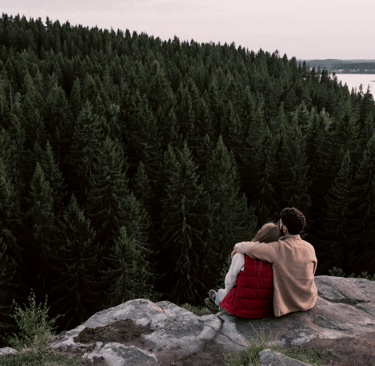 Couple sitting together on a cliff overlooking a pine forest, embracing each other
