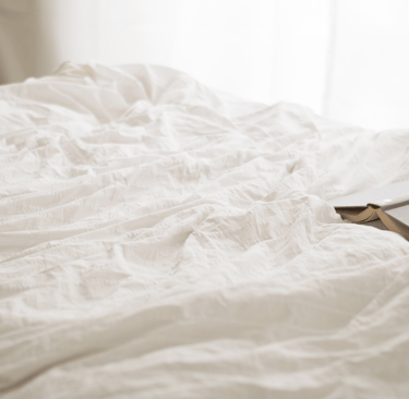 Unmade bed with soft white sheets and a closed journal on the mattress near a window.
