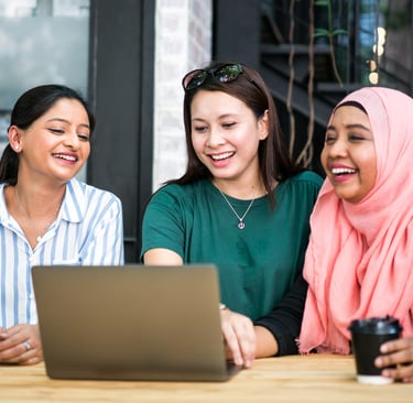 Three women smiling and looking at laptop while enjoying coffee outdoors