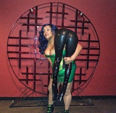 MsMadyson in a green latex dress carrying  a person over her shoulder who is wearing latex pants