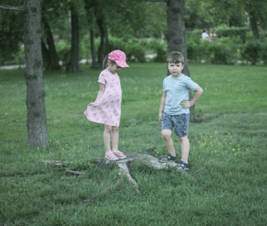 A boy and girl standing beside trees, exploring a wooded area during daytime.