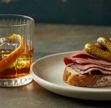 A close-up photograph featuring a classic Rum Old fashioned cocktail and a deli sandwich. 