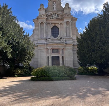 Historic stone chapel with classical architecture, green trees, and a gravel courtyard under a blue sky.