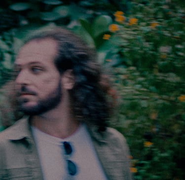 Candid portrait of a bearded man with long hair looking away against a blurred garden background.