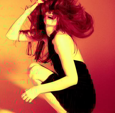 Fashion model in a black mini dress and high heels posing with wind-swept hair under warm orange lighting.