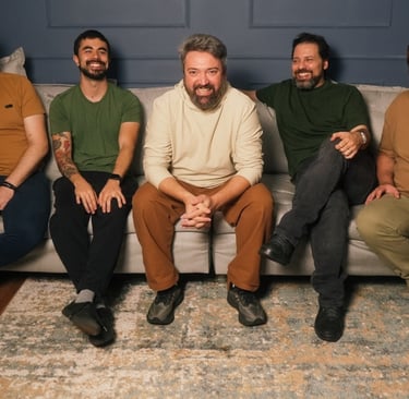 A smiling five-member rock band sitting on a grey sofa in a studio with drums visible in the background.