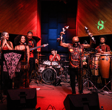 Live Afro-fusion band performing with conga drums, percussion, and singers in a recording studio.