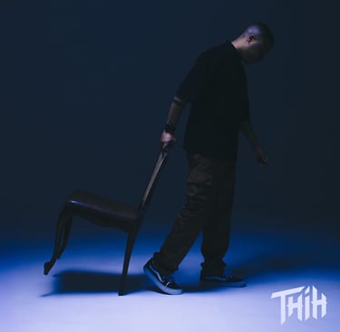A man in streetwear pulling a chair across a blue-lit studio floor for a moody photoshoot.