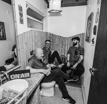 Three men talking in a bathroom-themed studio with an On Air sign and bamboo wall decor.