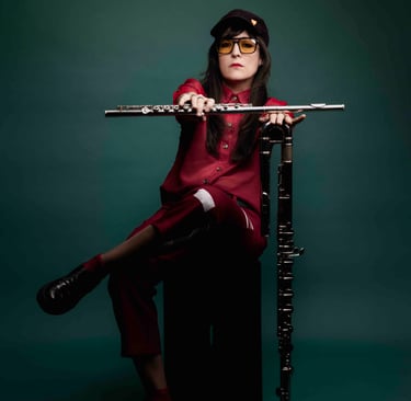 A female flutist in a red outfit holding a silver flute and standing bass flute against a dark green background.