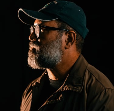 Side profile of a middle-aged man with a gray beard wearing a baseball cap and glasses against a dark background.