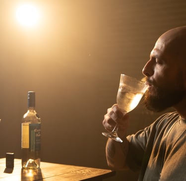A bearded man drinking white wine from a glass at a candlelit table with a bottle.