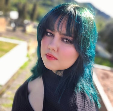 Young woman with blue and green dyed hair and bangs wearing makeup in a sunny outdoor setting.