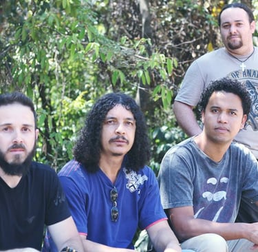 A four-member rock band with long hair and beards posing outdoors in front of green trees.