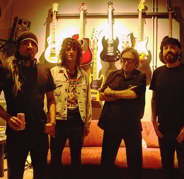 A four-member rock band stands in a music studio with electric guitars mounted on the wall behind them.