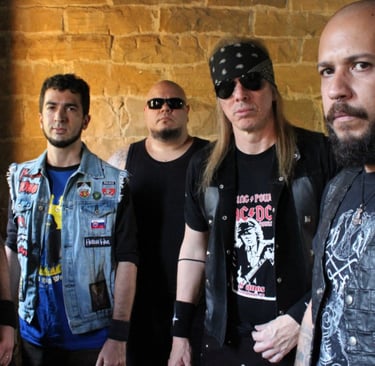 Heavy metal band members wearing denim vests and band t-shirts posing against a stone wall.