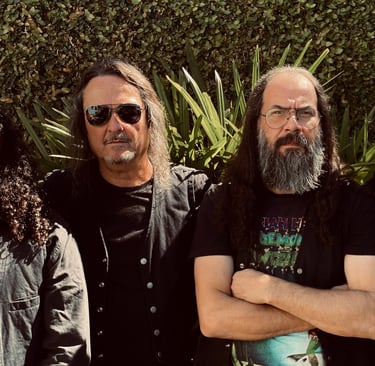 A four-member heavy metal rock band with long hair and beards posing outdoors against a green hedge.