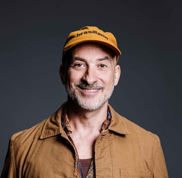 A smiling man with a beard wearing a yellow Brasiliano cap and a tan jacket against a dark background.
