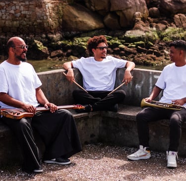 Three musicians holding instruments outdoors sitting on a stone bench near a rocky shoreline.