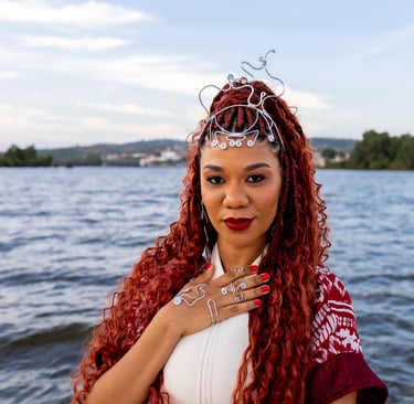 Woman with red braids and silver wire jewelry posing by a scenic river at sunset.