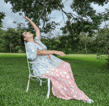 Woman in floral kaftan dress posing on white chair in a lush green garden.