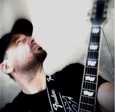 A man wearing a black baseball cap and t-shirt playing an electric guitar with a Fender strap.
