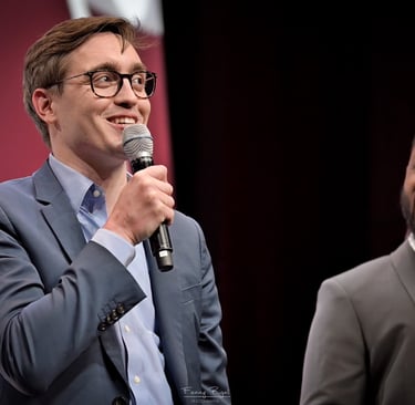 A smiling male speaker in a blue suit holding a microphone at a conference stage
