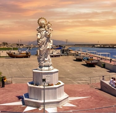 A white marble statue of the Virgin Mary overlooking a coastal fishing harbor at sunset.
