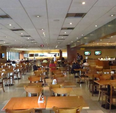 Spacious modern cafeteria with wooden tables and chairs, bright ceiling lighting, and large windows.