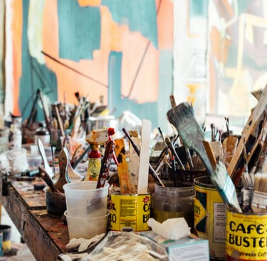 A collection of different sized used artist paintbrushes sit on a paint stained table.