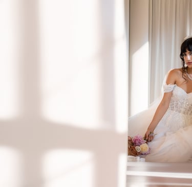 Sposa seduta in una stanza bianca, circondata da ombre geometriche di luce, con bouquet tra le mani