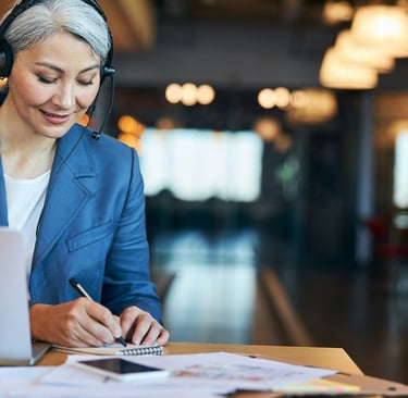 Businesswoman in headphones making notes while sitting at a desk with a laptop