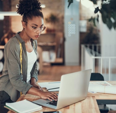 Young woman with a measuring tape typing on a notebook while standing near a desk with paperwork.