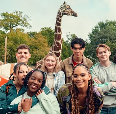 The Heartstopper ensemble cast posing in front of giraffe during season 3 filming.