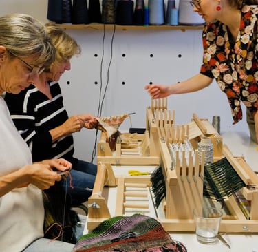 Weaving workshop instructor assisting students with small table looms and colorful yarn.
