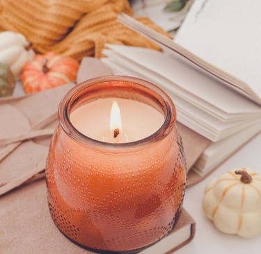 Lit orange candle in a textured glass jar, resting on suede notebooks with an open journal, mini pum