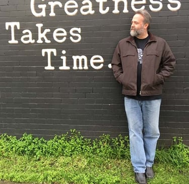 Jason leaning against a wall with a painted message that greatness takes time