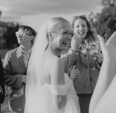 A joyful blonde bride laughs during her outdoor wedding reception with guests in a candid black and white photo.